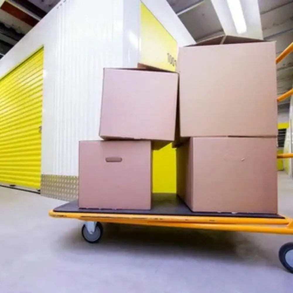 Stacked cardboard boxes on a moving trolley in front of a yellow storage unit. Convenient solution for loading and unloading items.