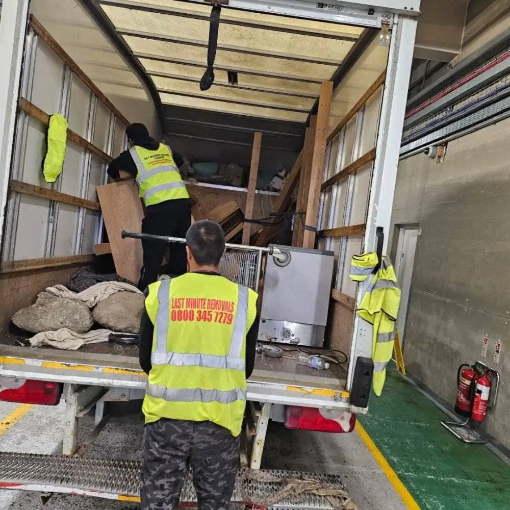 Two movers from Last Minute Removals organize various household items inside a packed moving truck parked in an indoor storage facility.